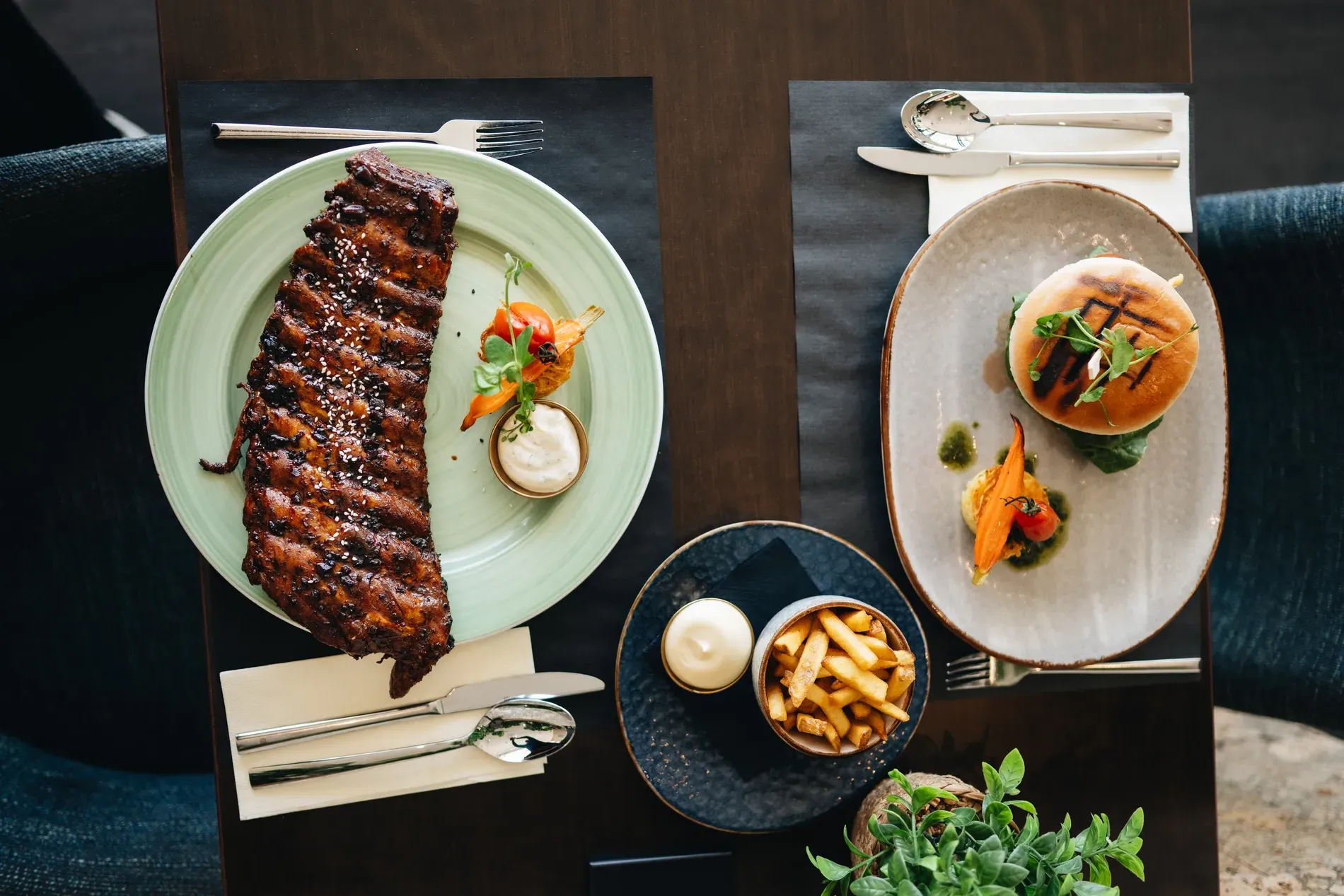 Horizontaal bovenaanzicht van een restauranttafel met een malse, grote streng spareribs op een groen bord, een hamburger op een grijs bord en een schaaltje frites met mayonaise in het midden.